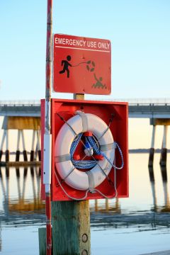 A life ring in a red emergency box is mounted on a wooden post next to water, with a red 